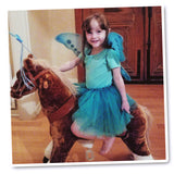 Little girl joyfully riding a toy horse, adorned in a whimsical turquoise green butterfly costume set, featuring wings, tutu, and wand. A delightful moment capturing the magic of imaginative play.
