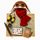 A styled flat-lay of a vintage aviator hat with round brown goggles, a chunky red knit scarf, yellow binoculars, a feather, a compass, a gemstone collection, a tree branch pencil, and a small notebook on a world map.