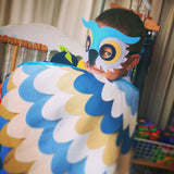 Boy wearing blue owl mask and cape, smiling and hiding behind raised wing
